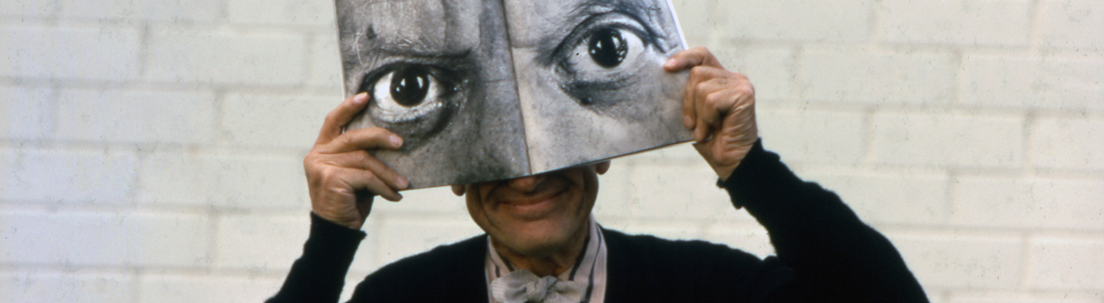 Charles Eames holds an open magazine to his face; on the two pages of the magazine are large men's eyes in black and white