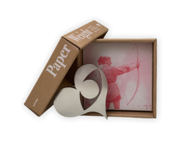 the Seeing with the Heart paper weight outside of its box. The lid is propped against the bottom with "paper weight" visible. Inside the box is a pink photo of Lucia Eames firing a bow.